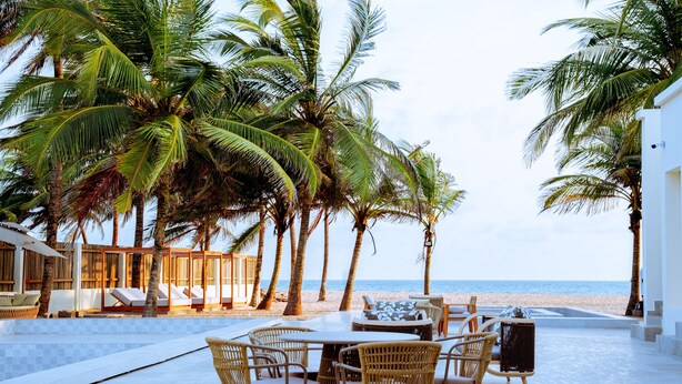 A poolside area with lounge chairs, a dining table, and chairs, surrounded by palm trees and a view of the beach. - Atlantic Shores Resort (Lagos)