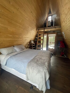 Ein Schlafzimmer einer Holzhütte mit einem Bett, einer Leiter, einem Fenster und einer Tür, die nach draußen führt.