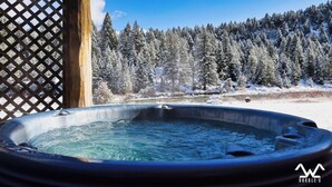 Ein Whirlpool in verschneiter Winterlandschaft mit Holzzaun und Wald im Hintergrund.