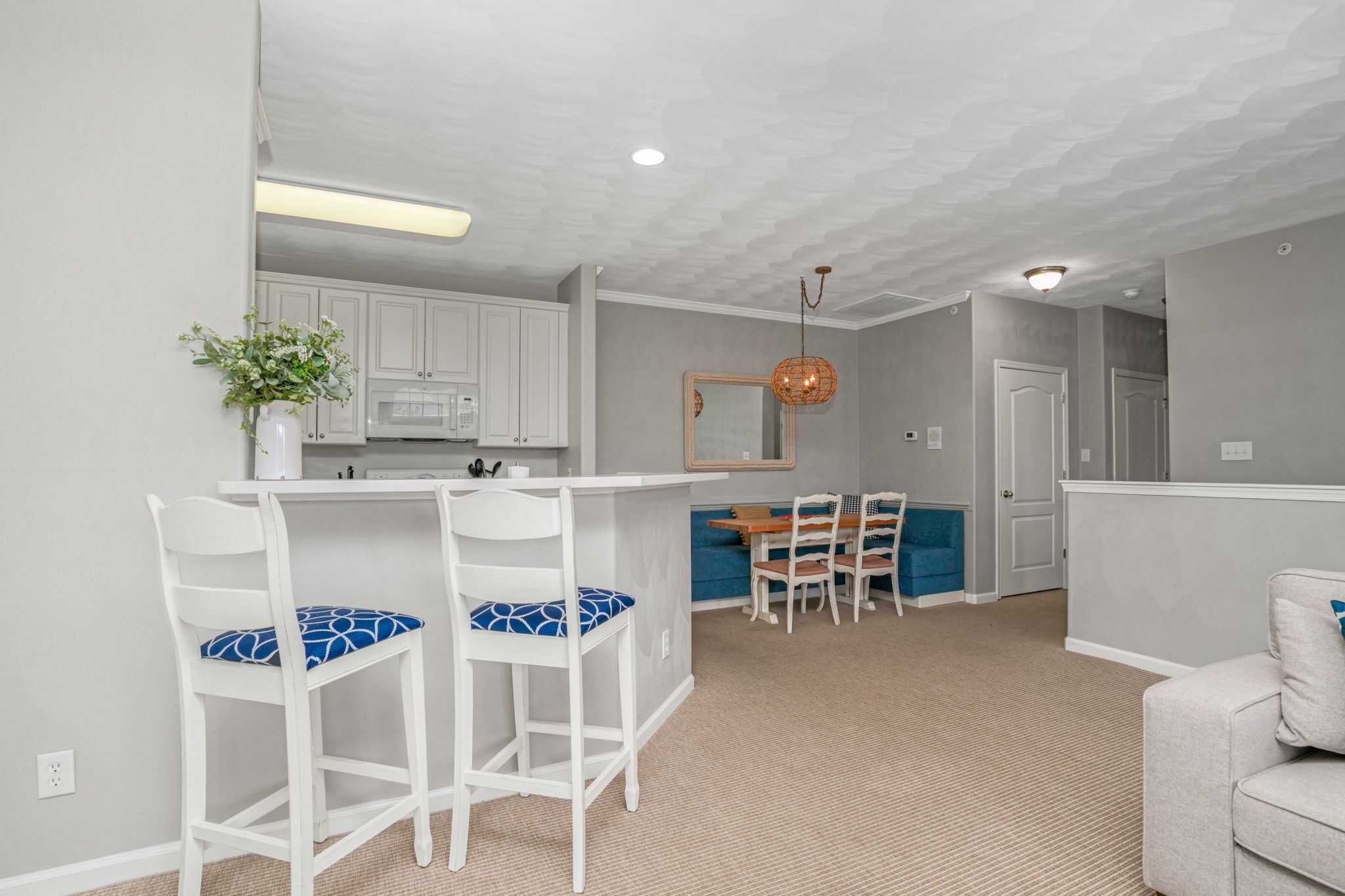 Une cuisine moderne avec des meubles blancs, un coin bar doté de deux tabourets de bar blancs, une table à manger avec des chaises et un luminaire suspendu.