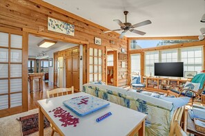A cozy cabin interior with wooden walls, a ceiling fan, a large TV, and a table with a board game. - Sunrise View | Waterfront, Kayaks, Gorgeous Sunrises! (Chincoteague Island)