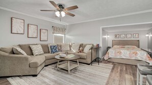 A living room with a beige sofa, a coffee table, and a ceiling fan. There are framed pictures on the wall and a bed with a floral bedspread in the background. - Grandpa's Cottage | Cute, clean and cozy studio apartment! (Chincoteague Island)