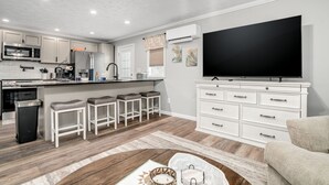A modern kitchen with white cabinets, a large TV, and bar stools. - Grandpa's Cottage | Cute, clean and cozy studio apartment! (Chincoteague Island)