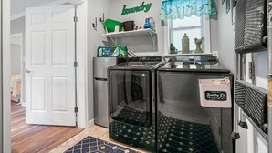 A laundry room with a washing machine, dryer, and shelves with laundry supplies. - Grandpa's Cottage | Cute, clean and cozy studio apartment! (Chincoteague Island)