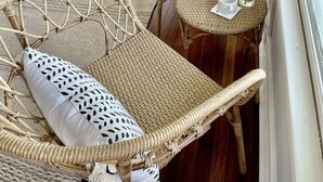 Un espace extérieur lumineux, aménagé pour se détendre, avec des meubles en osier et des coussins, une petite table ronde sur laquelle se trouvent une tasse et une bouteille, et une vue sur la verdure à travers de grandes fenêtres.