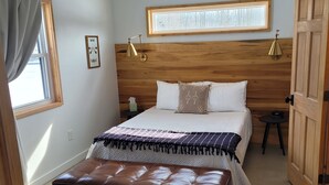 A bedroom with a wooden headboard, a large window, a bedside table, a brown leather bench, and a wooden door. - The Cork Cottages Unit 2, Arcade Table, 1K/1Q 2 Bath, New and Modern (Geneva)