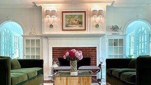 A spacious living room with a fireplace, two green sofas, a wooden coffee table with a vase of flowers, and a large chandelier.