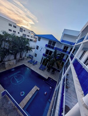 A swimming pool with lounge chairs, a palm tree, and a building with a blue roof. - Condo Neuf, Balcon en Plein Coeur de Sosua, 205 (Sosúa)