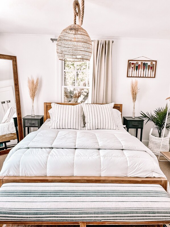A neatly arranged bedroom with a large bed, a hanging light fixture, a window with curtains, and a wall-mounted artwork.