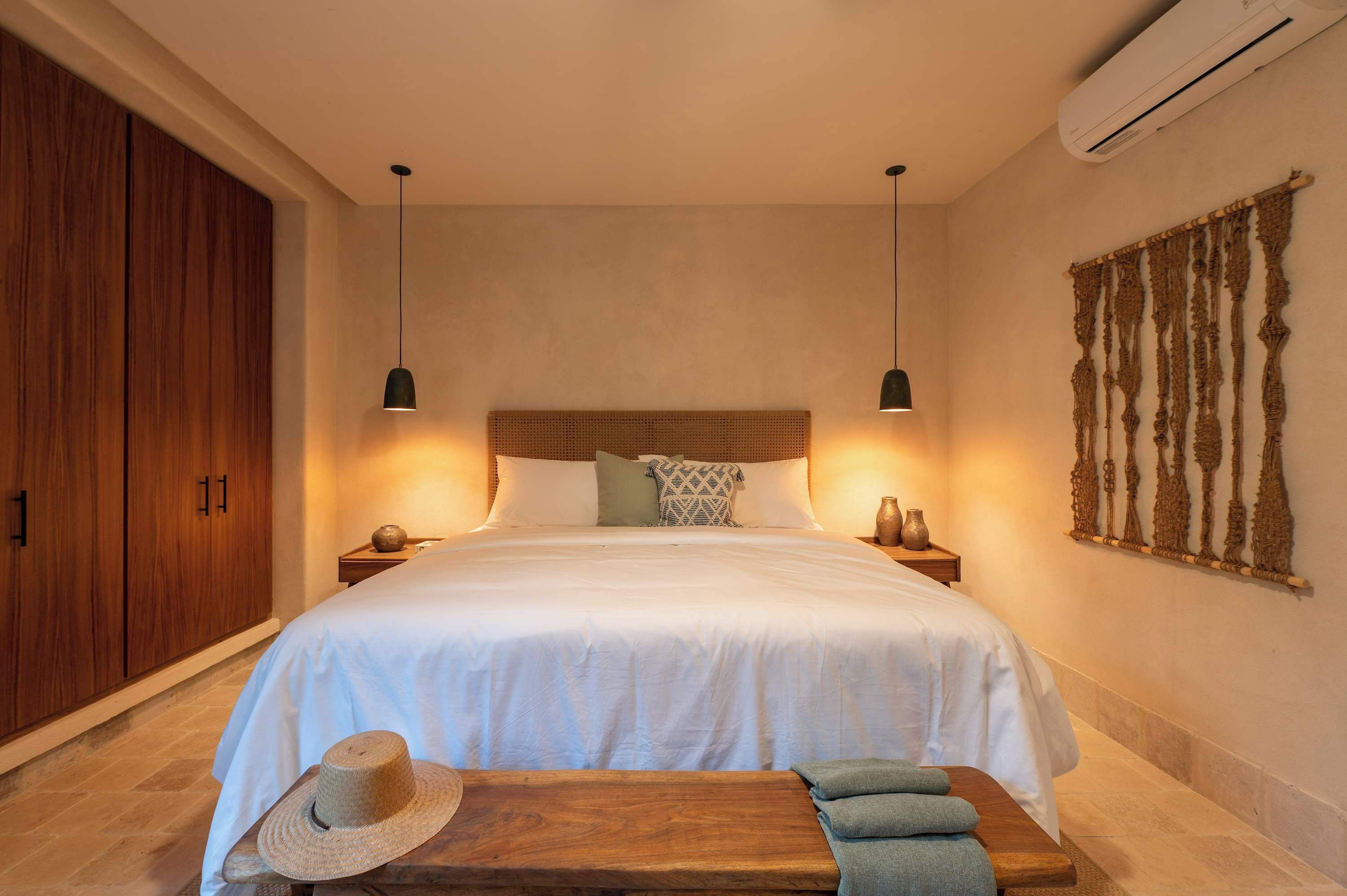A bedroom with a large bed, wooden wardrobe, two bedside tables, a wall-mounted air conditioner, and a decorative wall hanging.