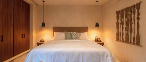 A bedroom with a large bed, wooden wardrobe, two bedside tables, a wall-mounted air conditioner, and a decorative wall hanging.