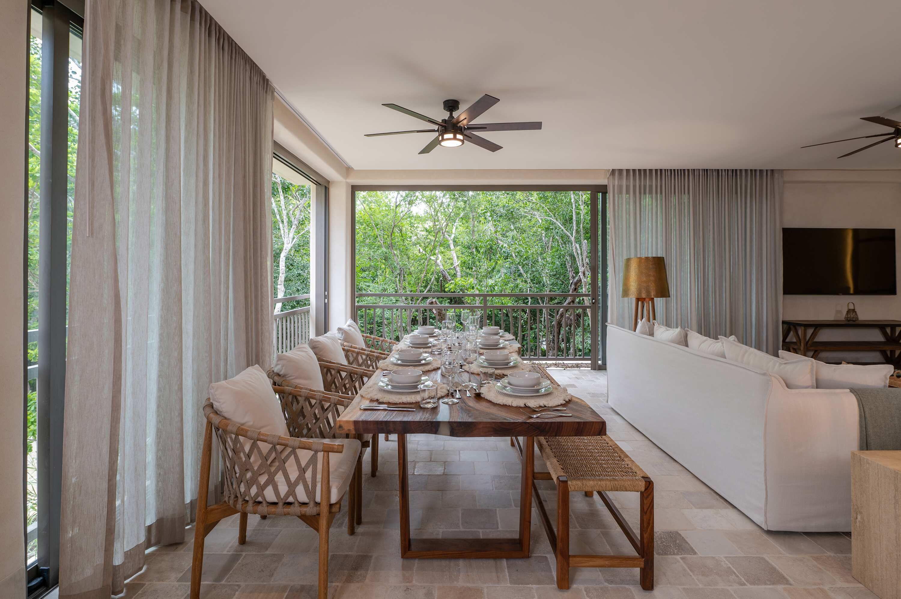 A dining area with a table set for four, a ceiling fan, and a balcony with a view of trees.