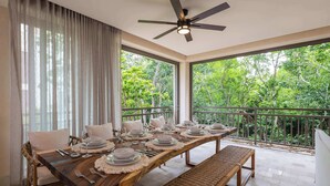 Un comedor con una larga mesa de madera dispuesta para una comida, ventanas amplias con vistas a zonas verdes y un ventilador de techo.