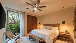 Un dormitorio moderno con una cama grande, un ventilador de techo, un banco de madera y vistas a zonas verdes a través de una puerta corredera.