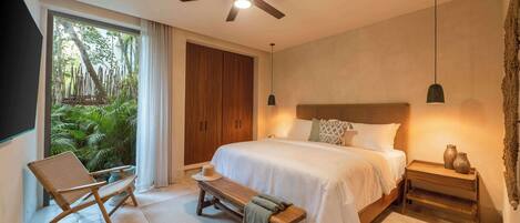 A modern bedroom with a large bed, a ceiling fan, a wooden bench, and a view of greenery through a sliding door.