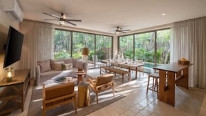 Amplia sala de estar con sofá, mesa de centro y mesa de comedor, ventanas amplias con vistas a exuberante vegetación.