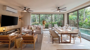 Amplia sala de estar con televisor de pantalla plana grande, asientos cómodos, mesa de comedor para cuatro personas y vistas a un jardín exuberante y piscina a través de amplios ventanales.