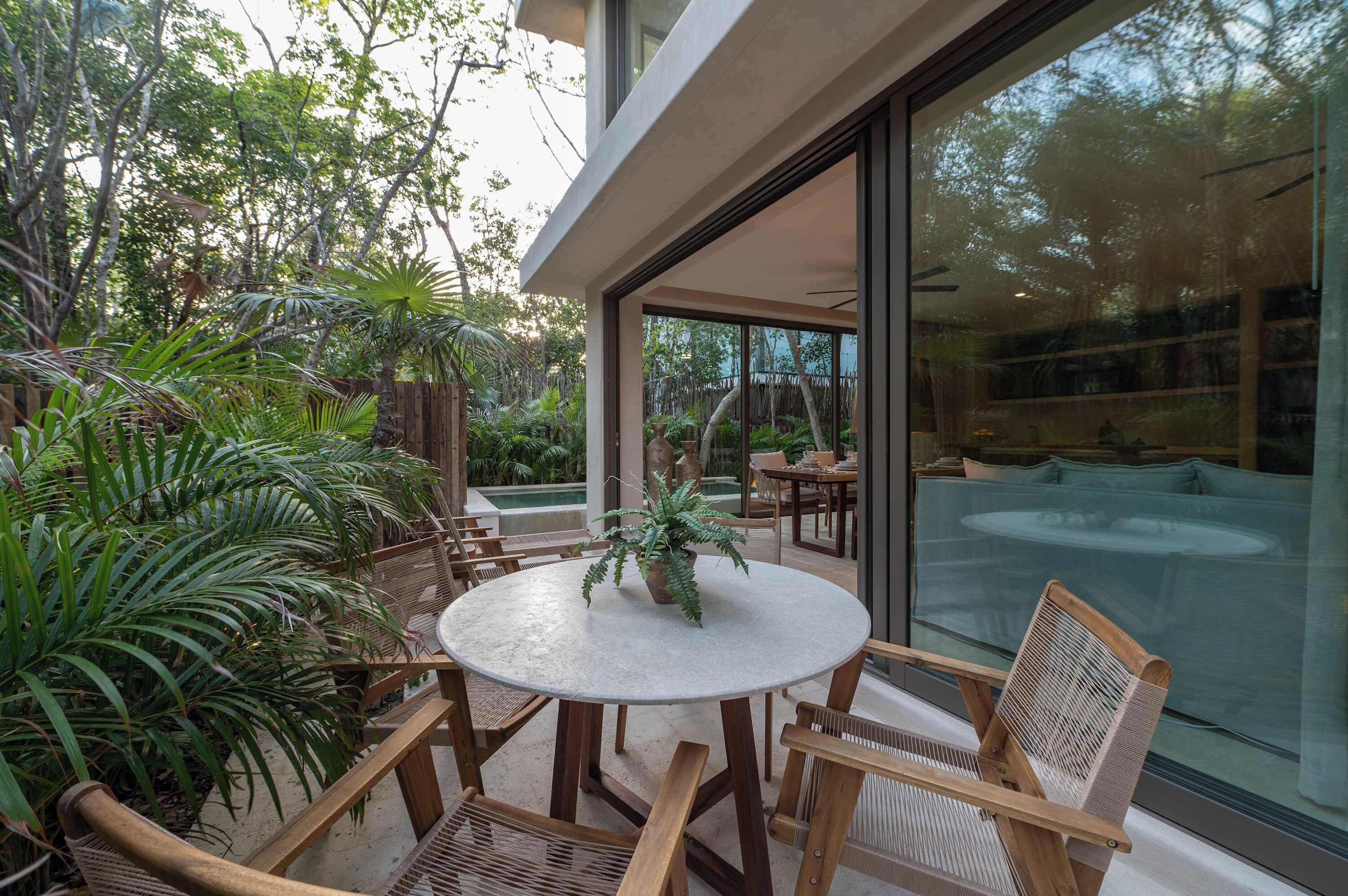 A modern outdoor seating area with a round table and wooden chairs, surrounded by lush greenery and a glass wall offering a view of the interior.