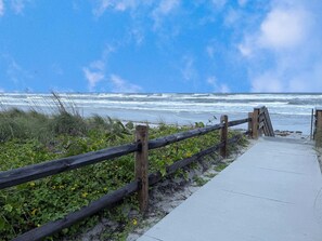 A coastal walkway with a wooden fence and a concrete path leading to the beach. - Pet-Friendly Coastal Retreat • Walk to Beach • Pools & Nature Trails (New Smyrna Beach)