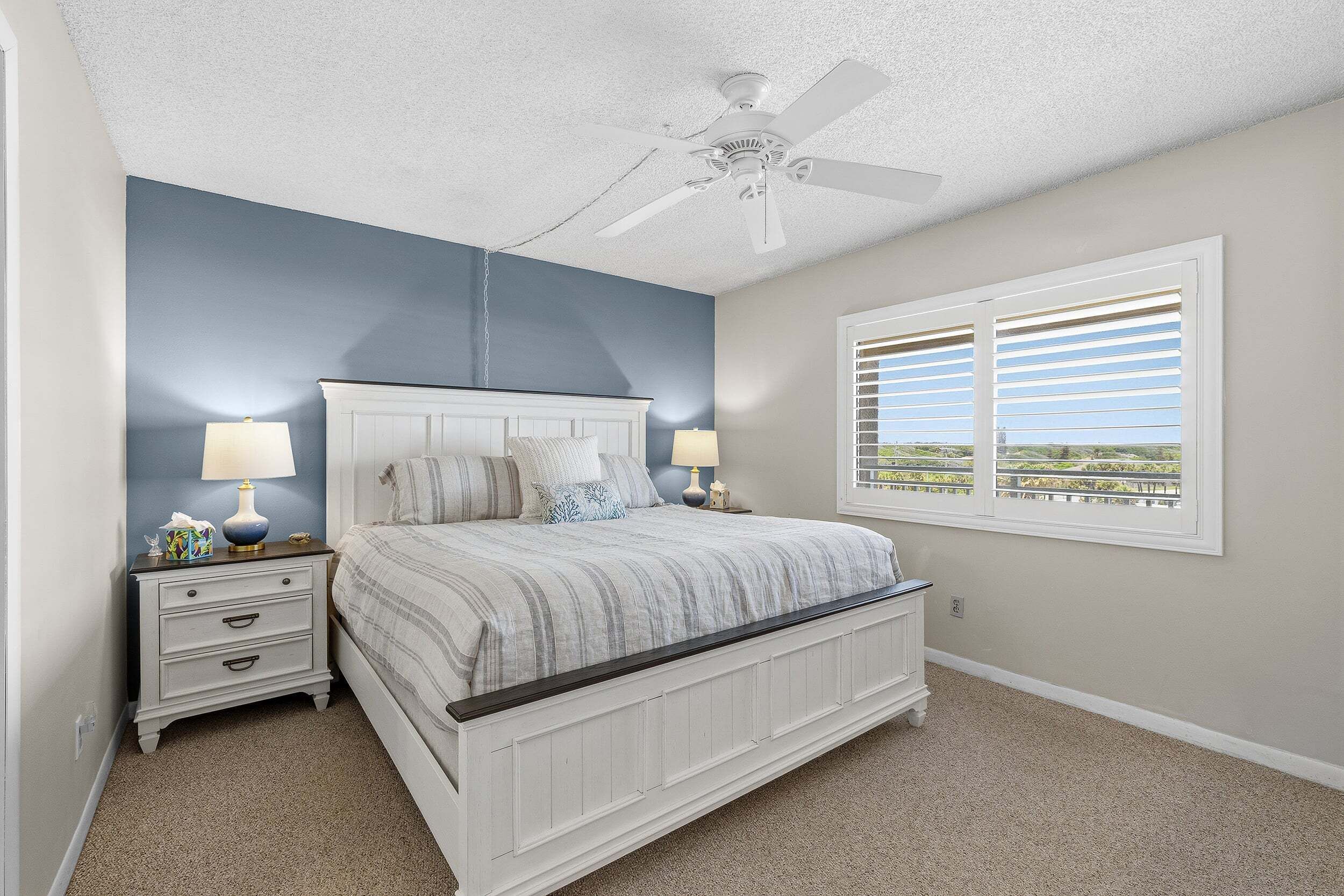 A bedroom with a bed, nightstands, lamps, a ceiling fan, and a window with blinds.