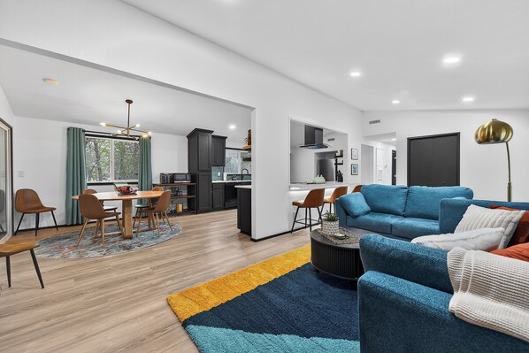 A modern living room with a blue sofa, a dining area with a wooden table, and a kitchen in the background.