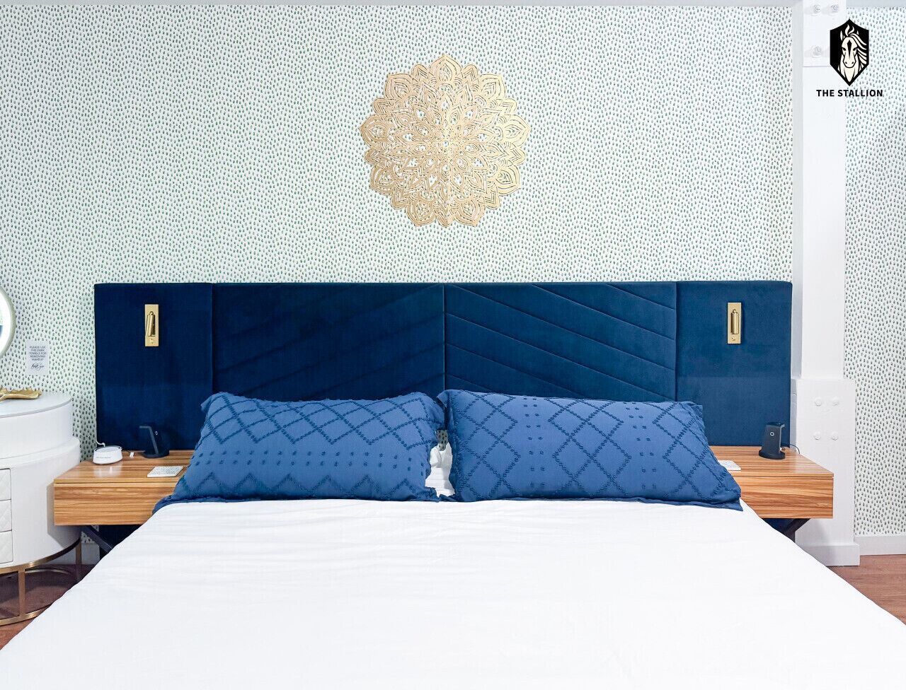A bedroom with a large bed, two nightstands, a decorative wall piece, and a logo in the top right corner.