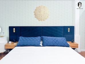 Una camera da letto con un letto grande, due comodini, un elemento decorativo a parete e un logo nell'angolo in alto a destra.
