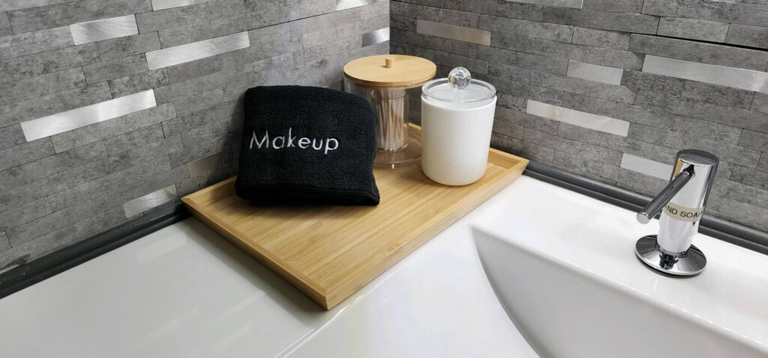 Un bagno con un vassoio di legno che contiene un asciugamano nero con l'etichetta 'Makeup', un contenitore bianco e un dispenser per sapone.