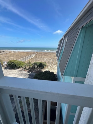 Blick von einem Balkon auf einen Strand mit Holzterrasse und einem Gebäude mit türkisblau verkleideter Fassade.