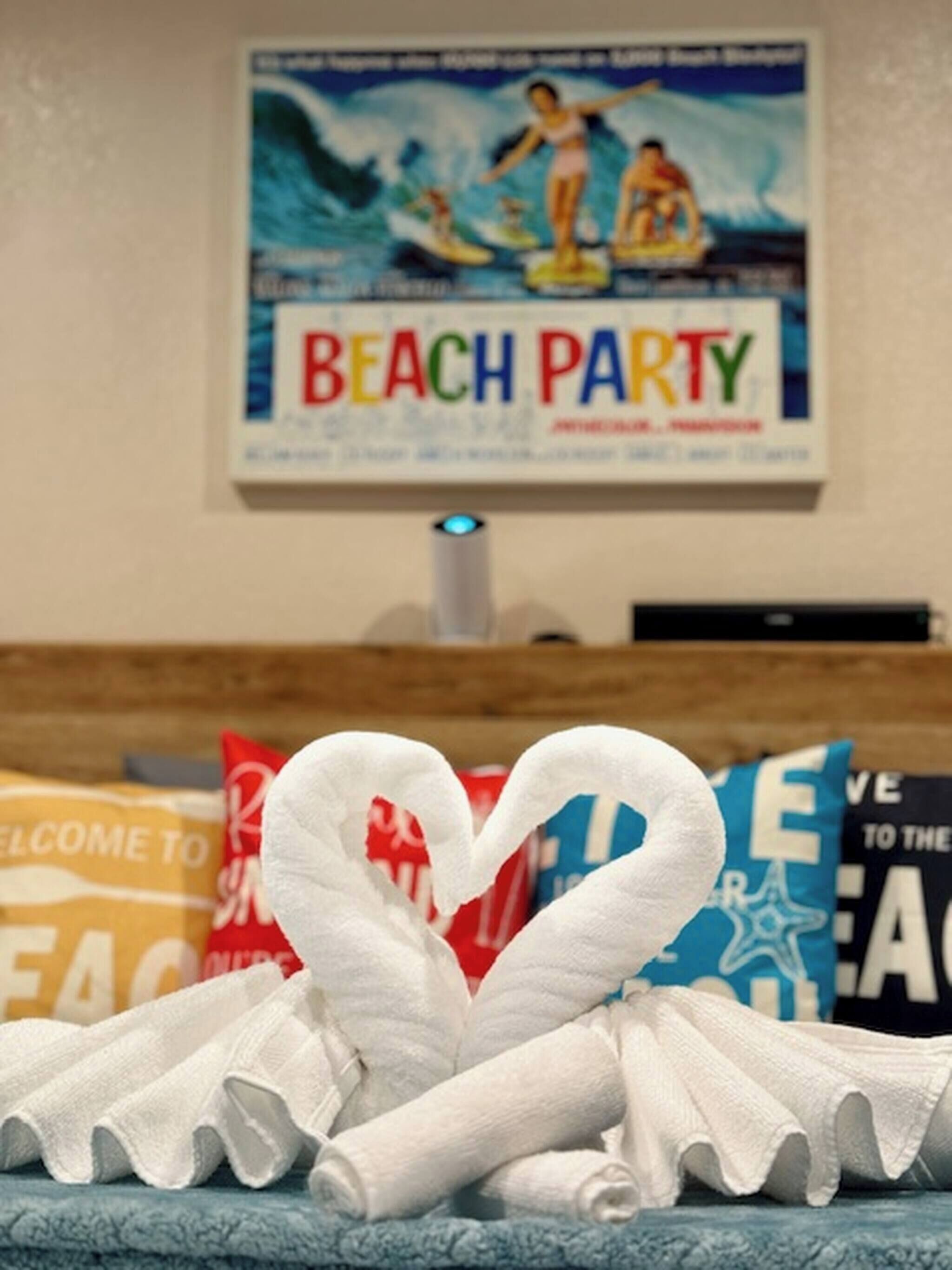 Een kamer met een handdoek in de vorm van een hart, een poster met de tekst 'Beach Party' aan de muur en kleurrijke kussens.