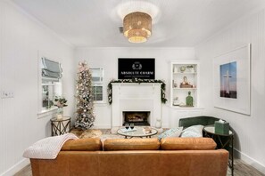 A cozy living room with a fireplace, a sofa, a coffee table, and a chandelier. - Songbird Cottage | 1/1, Mini Kitchen, Hot Tub, King Bed (Fredericksburg)