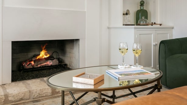 Habitació amb una llar de foc, una taula rodona de vidre amb un llibre i dues copes de vi, i un sofà verd.