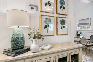 A room with a wooden table, a lamp, framed botanical prints, and a thermostat on the wall. - Songbird Cottage | 1/1, Mini Kitchen, Hot Tub, King Bed (Fredericksburg)