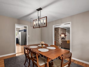 A dining area with a wooden table, six chairs, and a chandelier. There is a kitchen visible through an open doorway. - The North Asheville Ranch House; Upper Unit (Asheville)