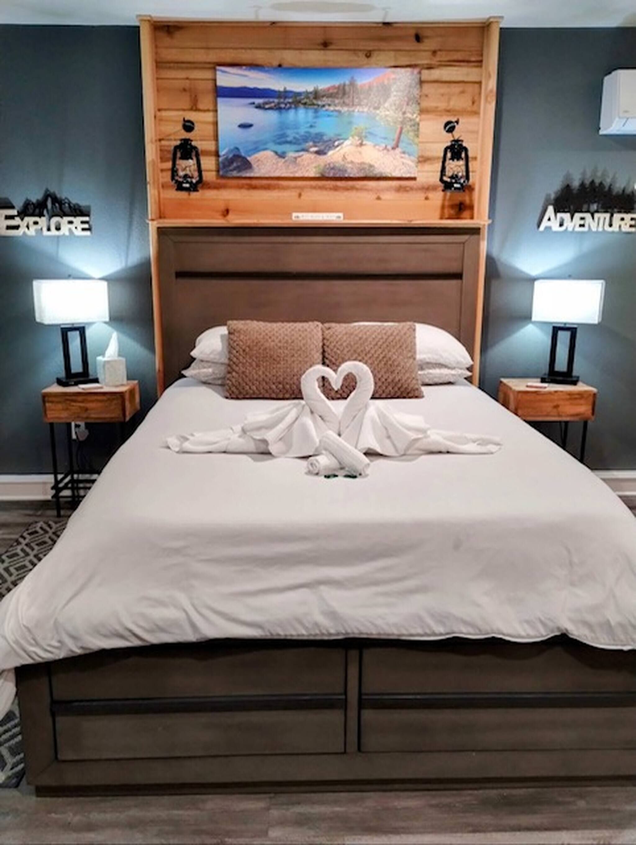 Een netjes opgemaakt bed met een handdoek in de vorm van een hart, een houten hoofdbord en twee bedlampjes.