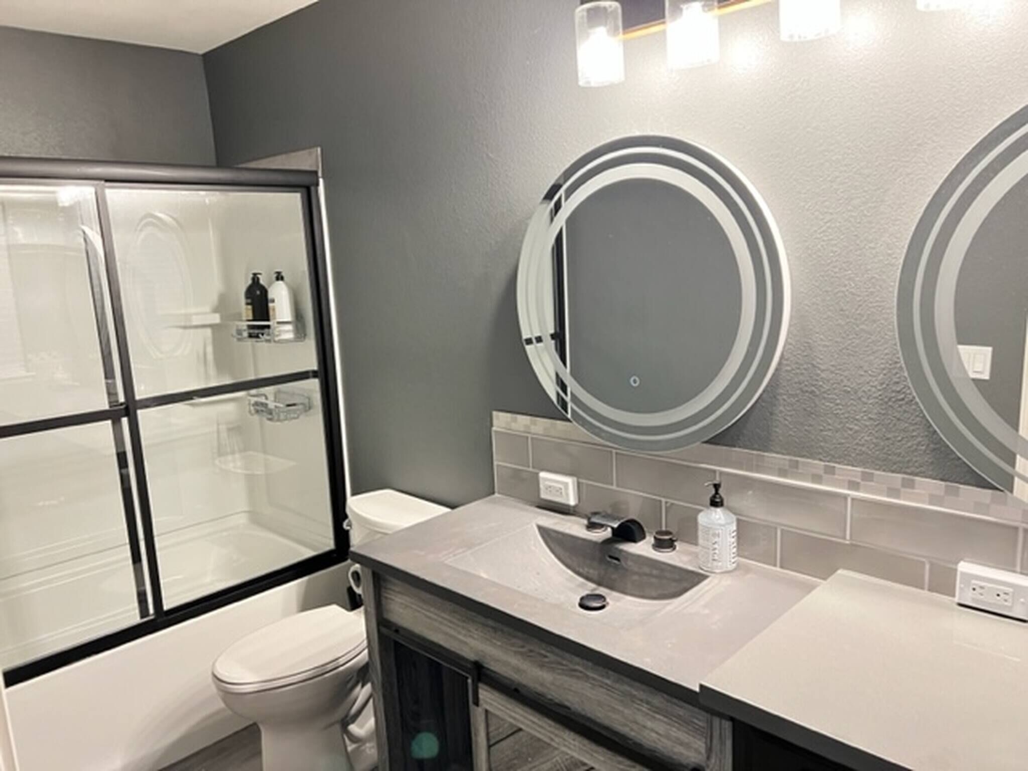 Un baño moderno con cabina de ducha de vidrio, inodoro, lavabo con espejo y dispensador de jabón.