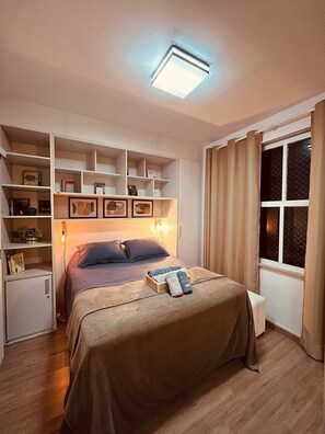 Ein Schlafzimmer mit einem Bett, einer eingebauten Regalanlage, einem kleinen weißen Schrank, einem Fenster mit Vorhängen und einer Deckenleuchte.