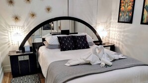 A modern bedroom with a curved headboard, a bed with white linens, a grey throw blanket, and a heart-shaped towel arrangement. There are two bedside tables with lamps, a patterned rug, and a wall with decorative star-like ceiling lights. - Deco Studio Retreat With Fireplace, Wifi, Queen Bed (Reno)