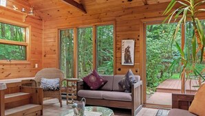 A wooden cabin interior with a sofa, wicker chairs, a glass coffee table, and large windows offering a view of the forest.