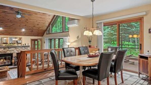 A spacious dining area with a large wooden table and black leather chairs, a stone fireplace, and large windows offering a view of the surrounding forest.