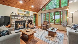 A cozy living room with a stone fireplace, wooden ceiling, and large windows offering a view of the forest.