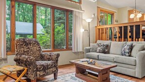 A living room with a patterned armchair, a sofa, a coffee table, and large windows offering a view of greenery.