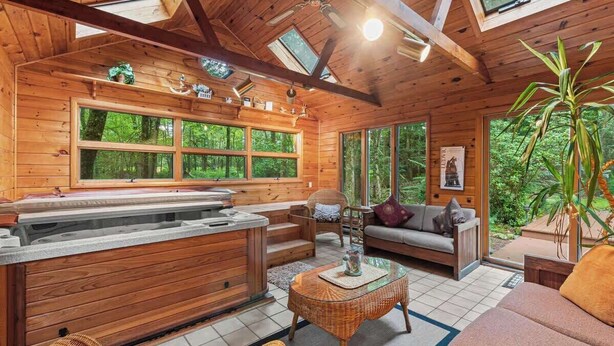 A wooden cabin interior with a hot tub, sofa, and large windows overlooking a forest.