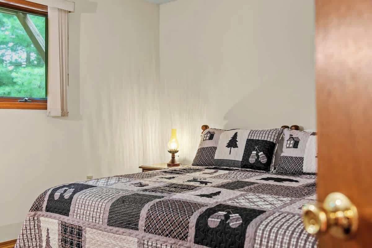 A bedroom with a bed covered in a quilt, a bedside table with a lamp, and a window with curtains.