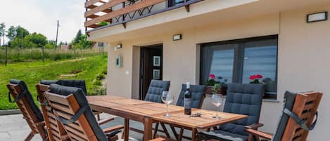 An outdoor patio with a wooden table and chairs, a balcony with a railing, and a view of greenery.