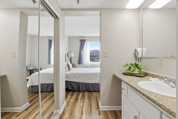 Ein Hotelzimmer mit Holzboden, einem Bett mit weißer Bettdecke, einem Badezimmer mit Waschbecken und Spiegel sowie Blick ins Freie.