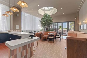 Eine moderne Hotelhalle mit einer zentralen Insellounge, Sitzbereichen und einem großen Fenster mit Blick ins Freie.