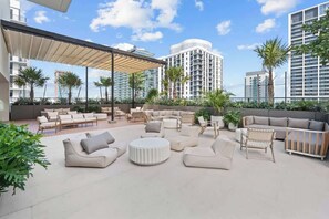 Eine Dachterrasse mit Außenbestuhlung, Palmen und modernen Gebäuden im Hintergrund.
