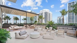 Un lounge en la azotea con mobiliario exterior, palmeras y edificios modernos al fondo.