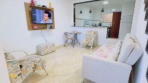 A living room with a TV, a sofa, a chair, a small table, and a kitchen in the background. - Deruci Beach House (Cabo Frio)